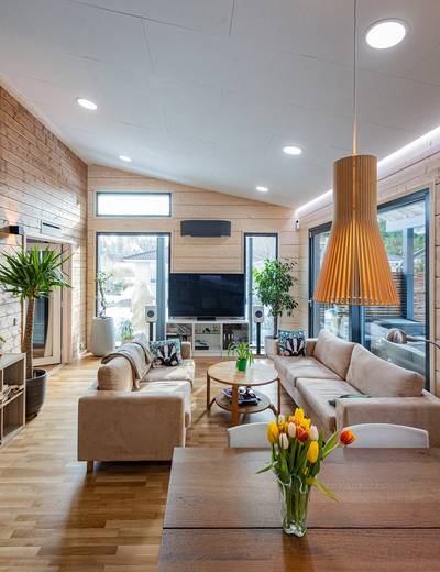Moderna sala de estar con grandes ventanales, sofás de color beige, TV, plantas, mesa de madera con jarrones tulipán, paredes de paneles de madera - iluminado con iluminación LED con estilo.