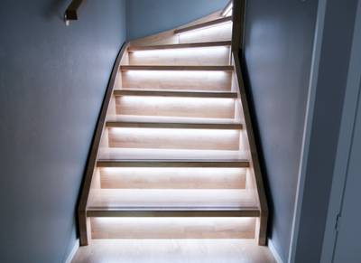 Una moderna escalera de madera con luces LED bajo cada peldaño, que resalta el diseño minimalista con una cálida iluminación sobre paredes grises.
