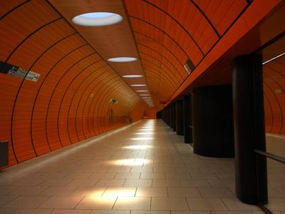 Unas luminosas lámparas LED de techo iluminan un moderno túnel de metro, cuyas paredes son de color naranja y el suelo está embaldosado.