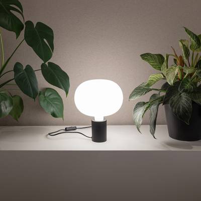 Moderna lámpara LED de sobremesa con pantalla redonda blanca sobre repisa, rodeada de macetas verdes para una iluminación elegante del hogar.
