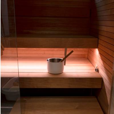 Interior de sauna de madera con cubo metálico y pala en los bancos; la cálida iluminación LED presenta los motivos de la madera en las superficies de asiento.