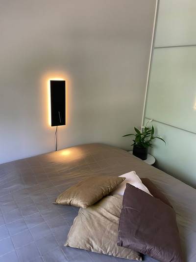 Un dormitorio acogedor con una cama bien hecha, cojines, un aplique LED retroiluminado y una planta en la mesilla de noche para darle un toque moderno.