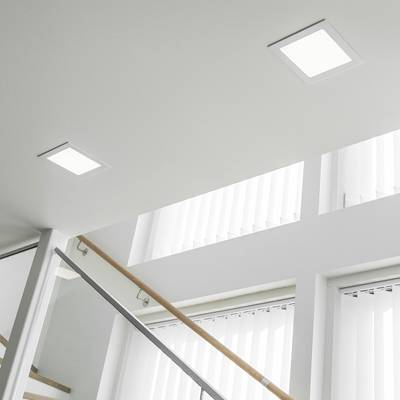 Unas brillantes luces cuadradas empotradas en el techo iluminan una moderna escalera con balaustrada de cristal y grandes ventanales con cortinas verticales.