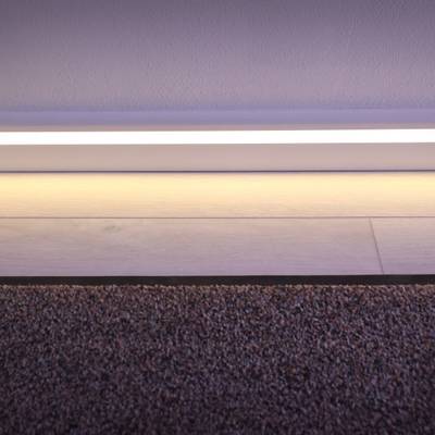 Una moderna tira de luz LED ilumina la transición entre la moqueta y el suelo de madera, resaltando los detalles blancos del zócalo.