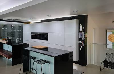 Cocina moderna con armarios blancos brillantes, encimeras negras, isla con taburetes de bar, horno doble de acero inoxidable, nevera con imanes justo debajo de la iluminación LED.