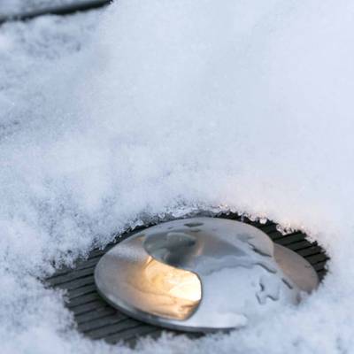 Texto alternativo: "Lámpara de suelo parcialmente cubierta de nieve que crea un cálido resplandor en el paisaje invernal - un escaparate de productos de la empresa de iluminación LED.