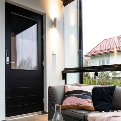 Moderna terraza acristalada con iluminación LED de bajo consumo, puerta negra, sofá gris con cojines de colores, paredes de cristal que dejan ver la casa con un vibrante tejado rojo.