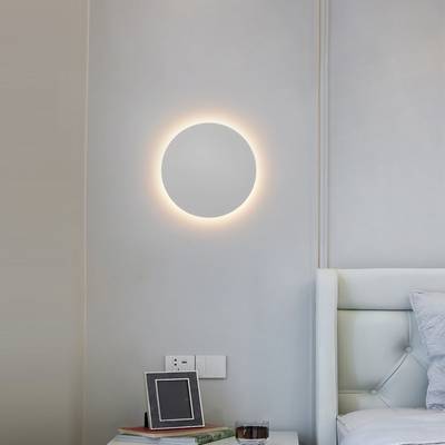 Un moderno aplique circular ilumina un dormitorio minimalista con paredes gris claro, un cabecero de cama blanco empenachado y una elegante decoración de mesilla de noche. Tienda de iluminación LED.