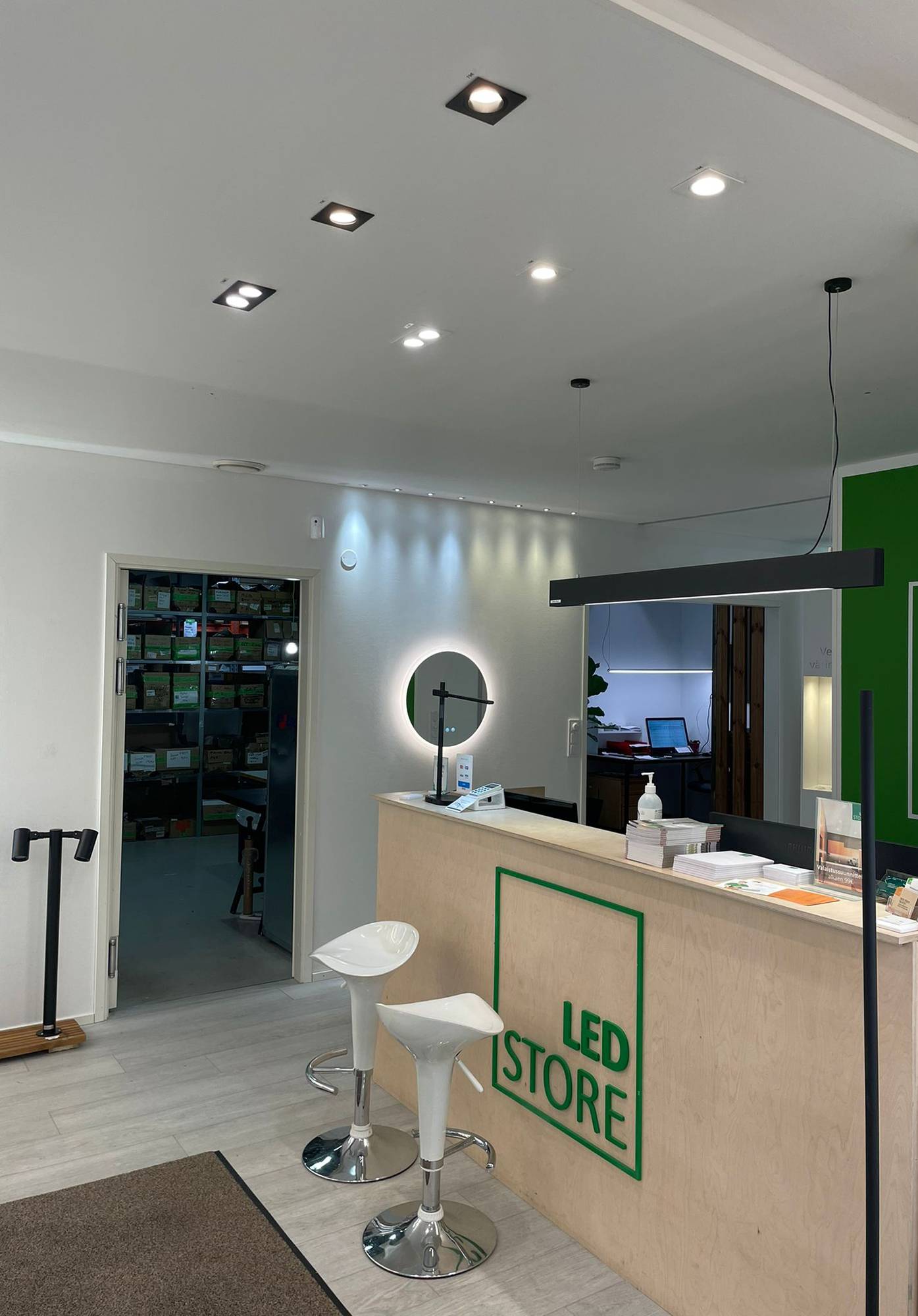 Luces led en la sala de exposición de Ledstore. Focos en el techo delante del mostrador y un plafón VIIVA encima del mostrador. La pared está acentuada por conjuntos de focos de 3W, fijos y direccionales. Espejo de luz Moon 600 bajo los focos. Ledstore.fi