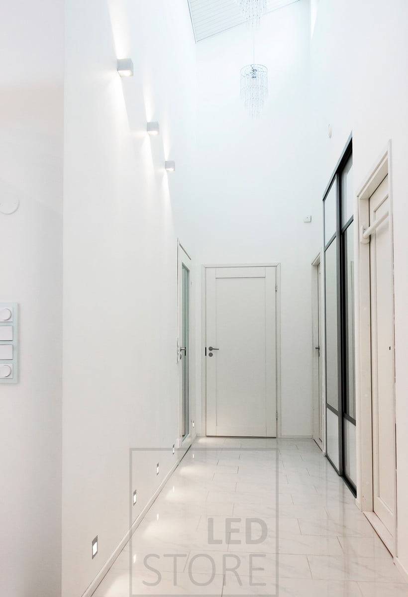 CUBIC luminaria led de pared que ilumina en dos direcciones en un pasillo para resaltar la altura del espacio. Los apliques led empotrados en el borde del suelo proporcionan luz indirecta al suelo. Ledstore.fi