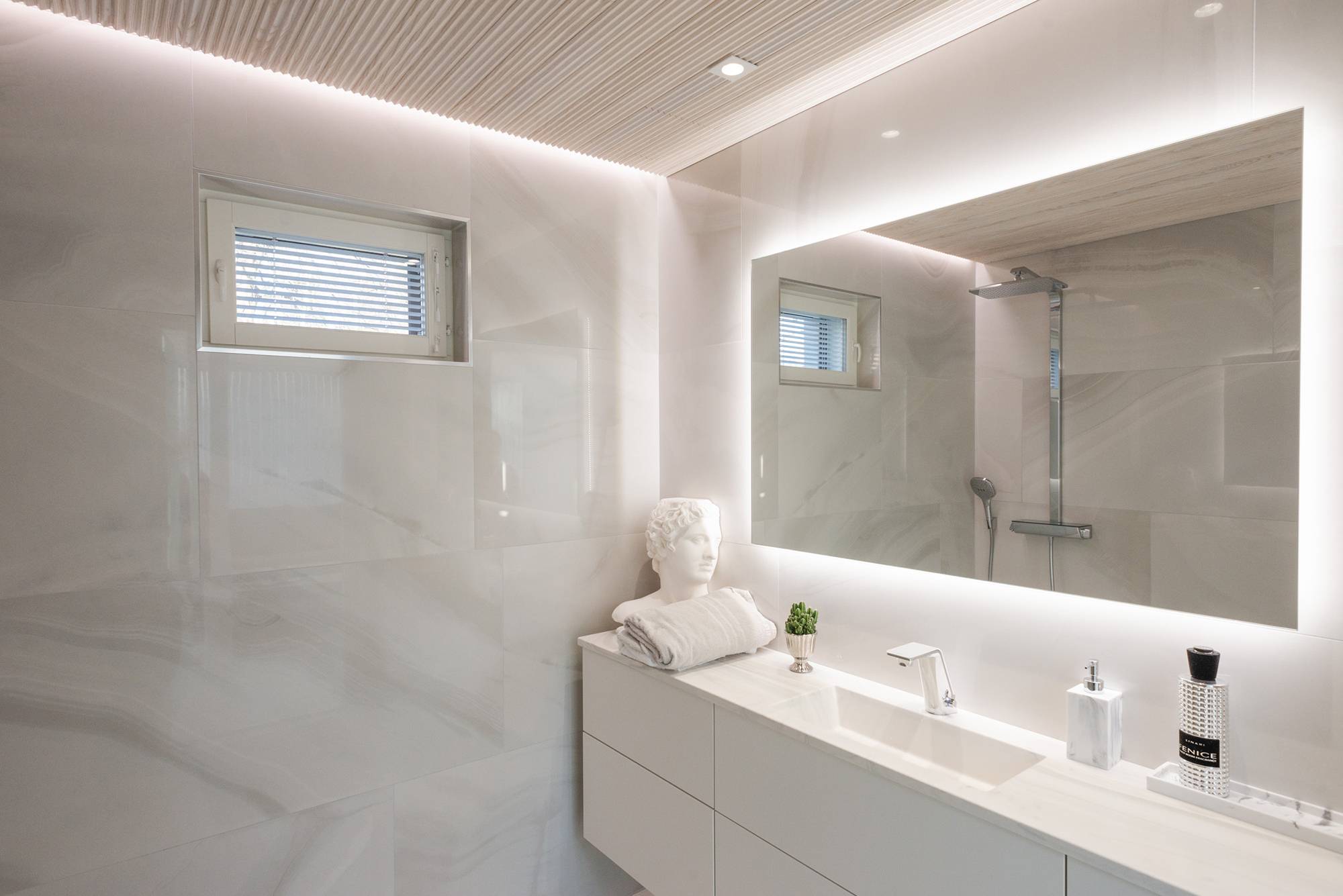 En un cuarto de baño con bañera de hidromasaje, un espejo de luz grande y espacioso combinado con focos y una tira de LED de pared a techo.