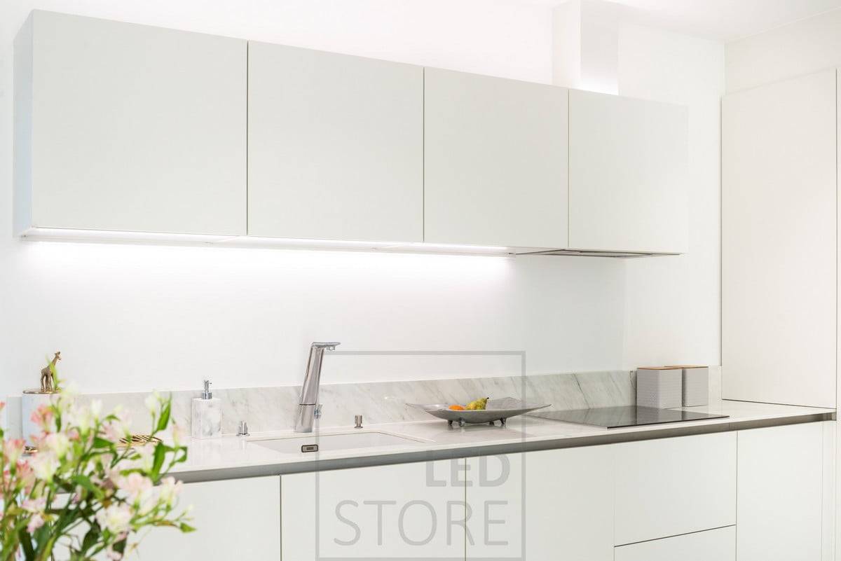 Tiras de LED encima de los armarios e iluminando la encimera. La luz neutra resalta los tonos limpios de la cocina blanca. Ledstore.fi