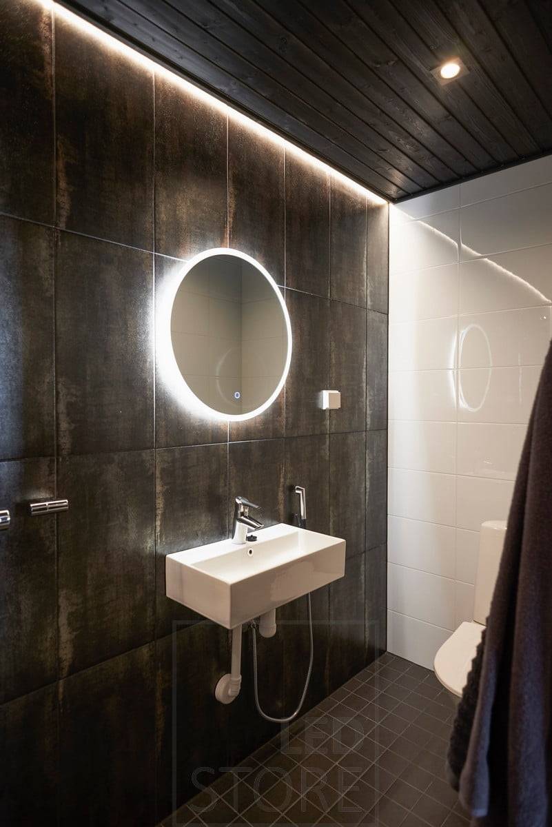Baño oscuro atmosféricamente iluminado con focos y luz indirecta. El espejo con luz tiene una función de desempañado que mantiene el espejo iluminado al salir de la sauna o de la ducha. Ledstore.fi