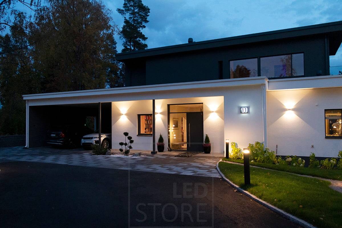 Aplique FUNK blanco bidireccional que ilumina la fachada y la entrada. La entrada está iluminada por luminarias de columna. Ledstore.fi