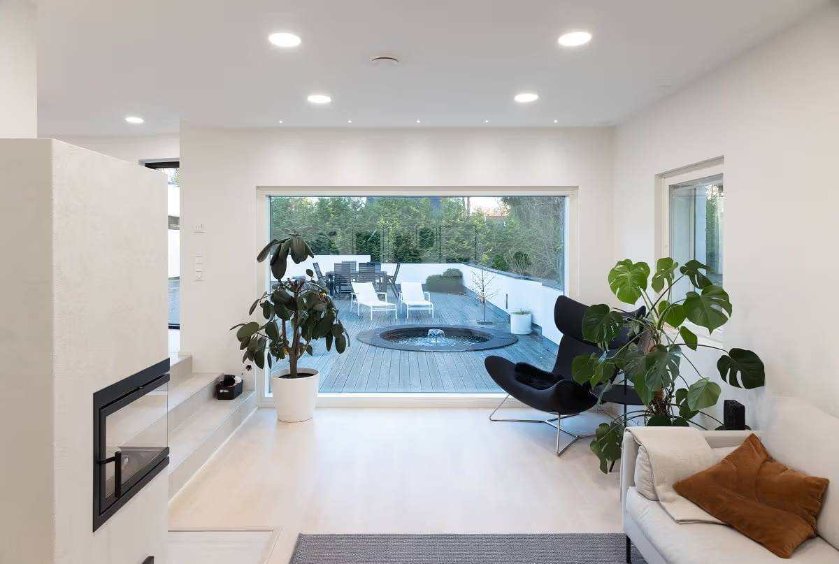 Un salón con una superficie luminosa y un gran ventanal con vistas al exterior. Hay paneles LED redondos en el techo y pequeños focos delante de la ventana.