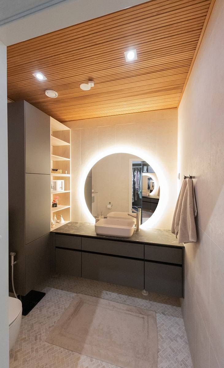 Un baño moderno con iluminación LED de LedStore.fi: un espejo circular retroiluminado con LED sobre el lavabo crea un ambiente elegante. El cuarto de baño tiene armarios oscuros, techos de madera cálida y paredes claras. Las toallas están colocadas a la derecha y las estanterías te esperan a la izquierda. En el suelo descansa una alfombra beige, que contribuye al efecto general.   