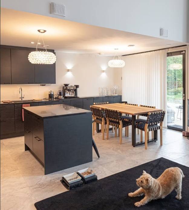 La moderna cocina y el comedor, con armarios negros, mesa de comedor de madera y una cómoda iluminación LED de LedStore.fi, crean un ambiente acogedor. Un gato estirado sobre una alfombra negra observa el acogedor conjunto.