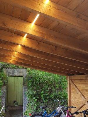Marquesina de madera, iluminada por una tira de LED que discurre entre el tejado y los parapetos. Bicicletas bajo la marquesina y una puerta verde al fondo.