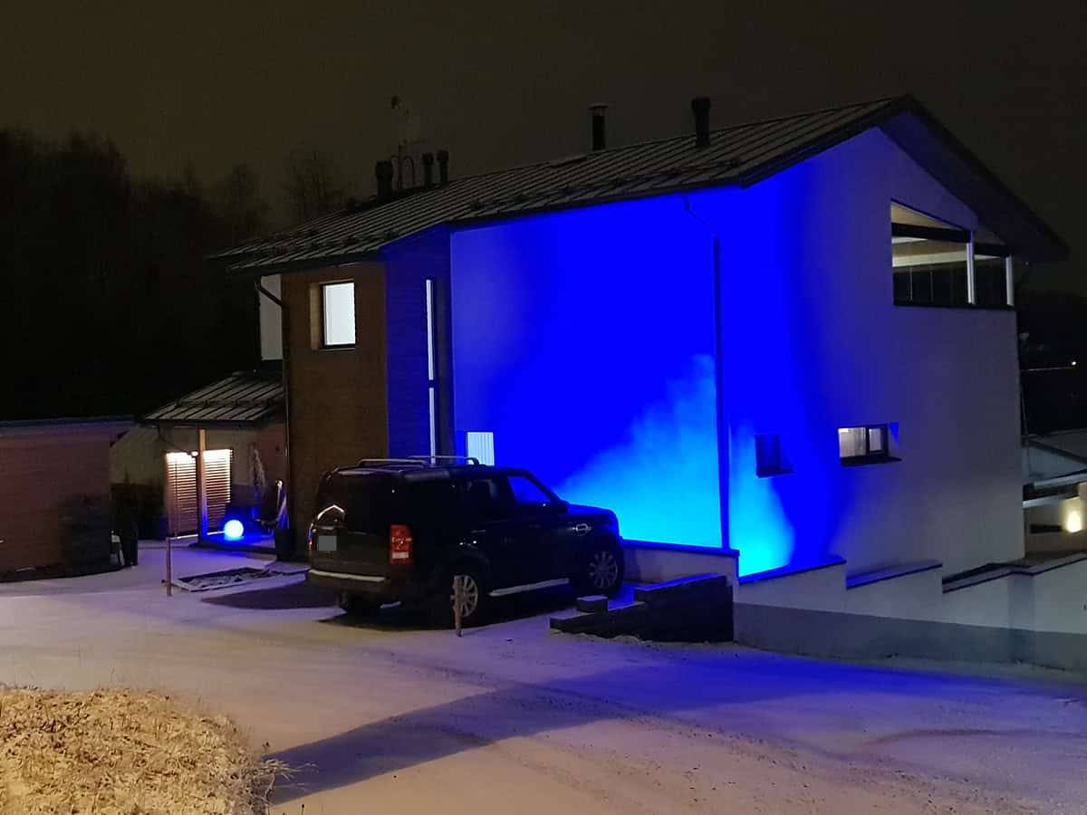 La fachada de una casa moderna iluminada de noche por luces LED azules. Un coche en el patio y una ligera capa de nieve en el suelo.