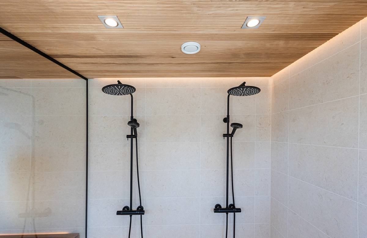 El moderno cuarto de ducha tiene dos rociadores negros para el alicatado de color crema. El techo de paneles de madera, acentuado por luces LED empotradas de LedStore.fi, crea un aspecto minimalista y elegante. La zona de ducha está separada del resto del espacio por una pared exterior de cristal.  
