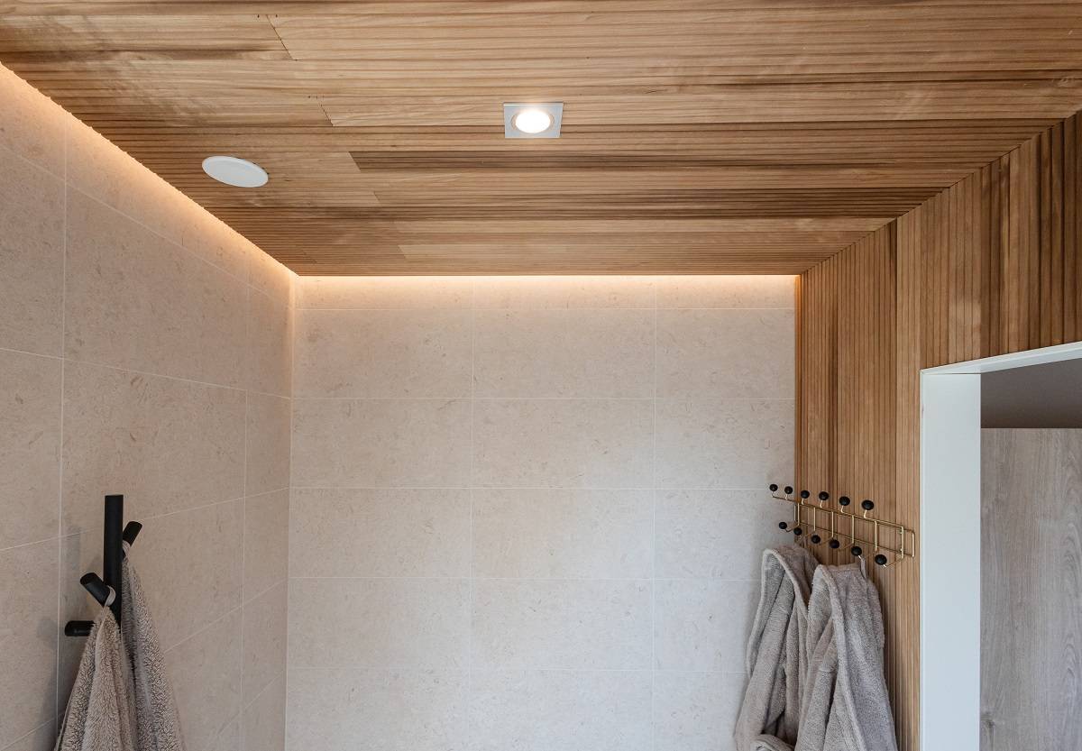 LedStore.fi ofrece una solución para el cuarto de baño moderno con una moderna iluminación LED. Las paredes de azulejos beige, el techo de madera y la iluminación LED empotrada crean un aspecto fresco y moderno. Las suaves toallas grises colgadas en ganchos negros armonizan el interior minimalista.