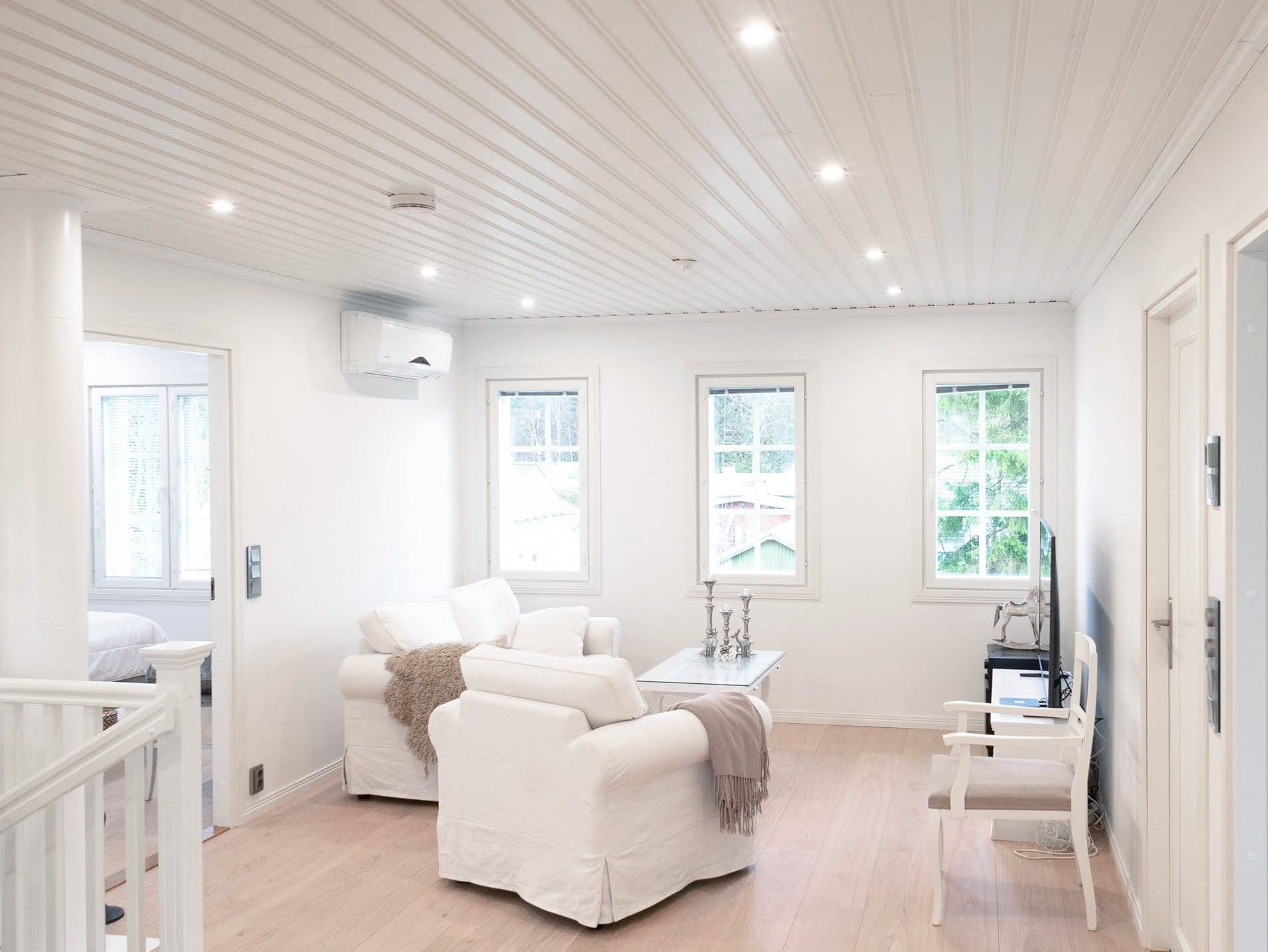 Moderno salón con iluminación LED, sillones blancos, mesa de centro y TV; entra más luz natural por los grandes ventanales y acentúa el suelo de madera.