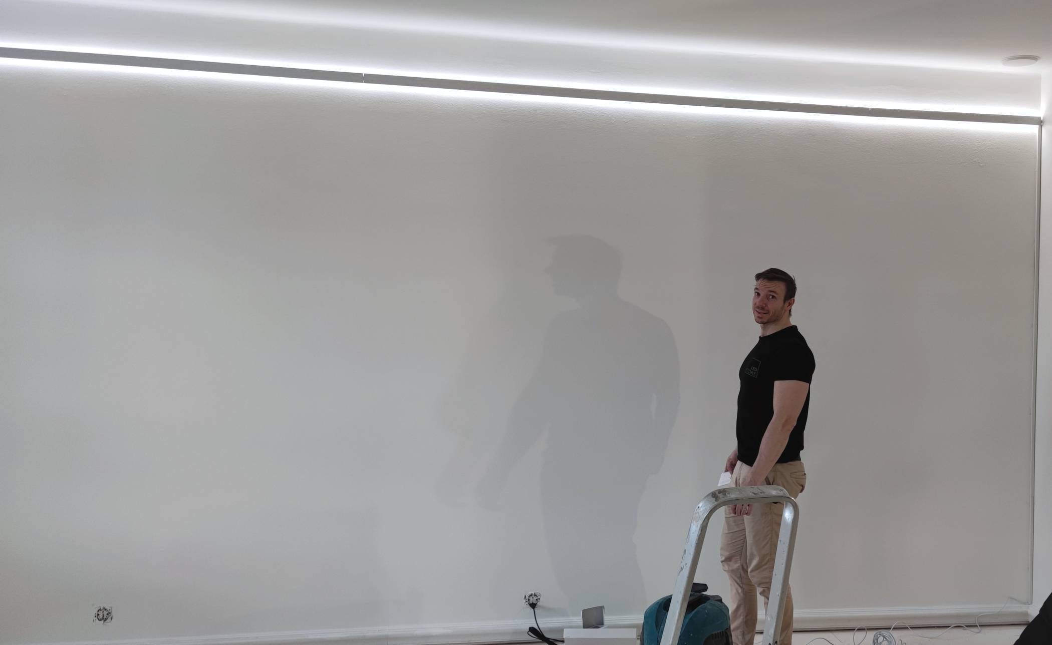 Una persona en una escalera con una herramienta, con iluminación LED suave desde arriba, contra una pared blanca. Cables y equipos esparcidos por el suelo con iluminación LED de taller.
