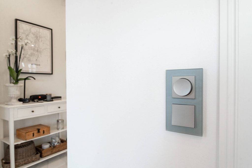 Moderno interruptor de luz LED con dos botones en una pared blanca; consola blanca con orquídea en maceta y mapa enmarcado en una habitación minimalista.