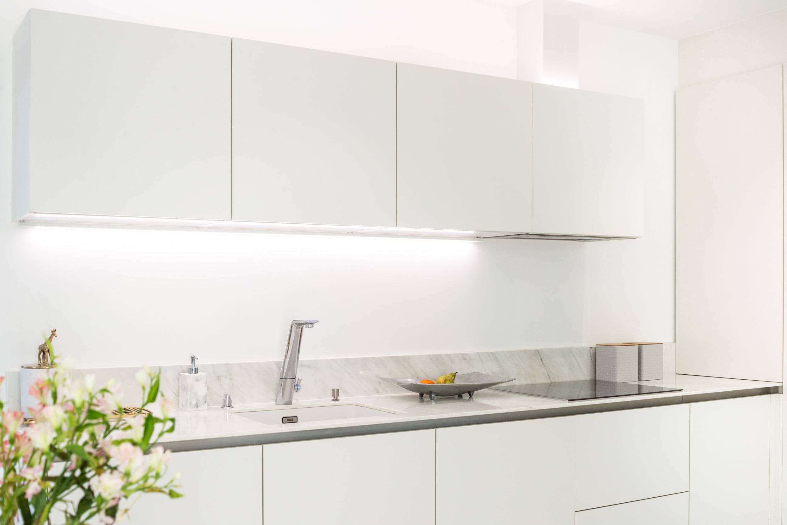 Tiras led para la cocina - ¡cómo elegir la iluminación para tu espacio! Una cocina elegante con armarios blancos limpios, moderna encimera de mármol y grifo de acero inoxidable. La iluminación LED bajo encimera acentúa el aspecto del espacio de trabajo. Una planta decorativa y un frutero añaden más estilo a la encimera. Puedes encontrar las soluciones de iluminación LED adecuadas en LedStore.fi, donde encontrarás productos LED de calidad para completar el aspecto de tu cocina.