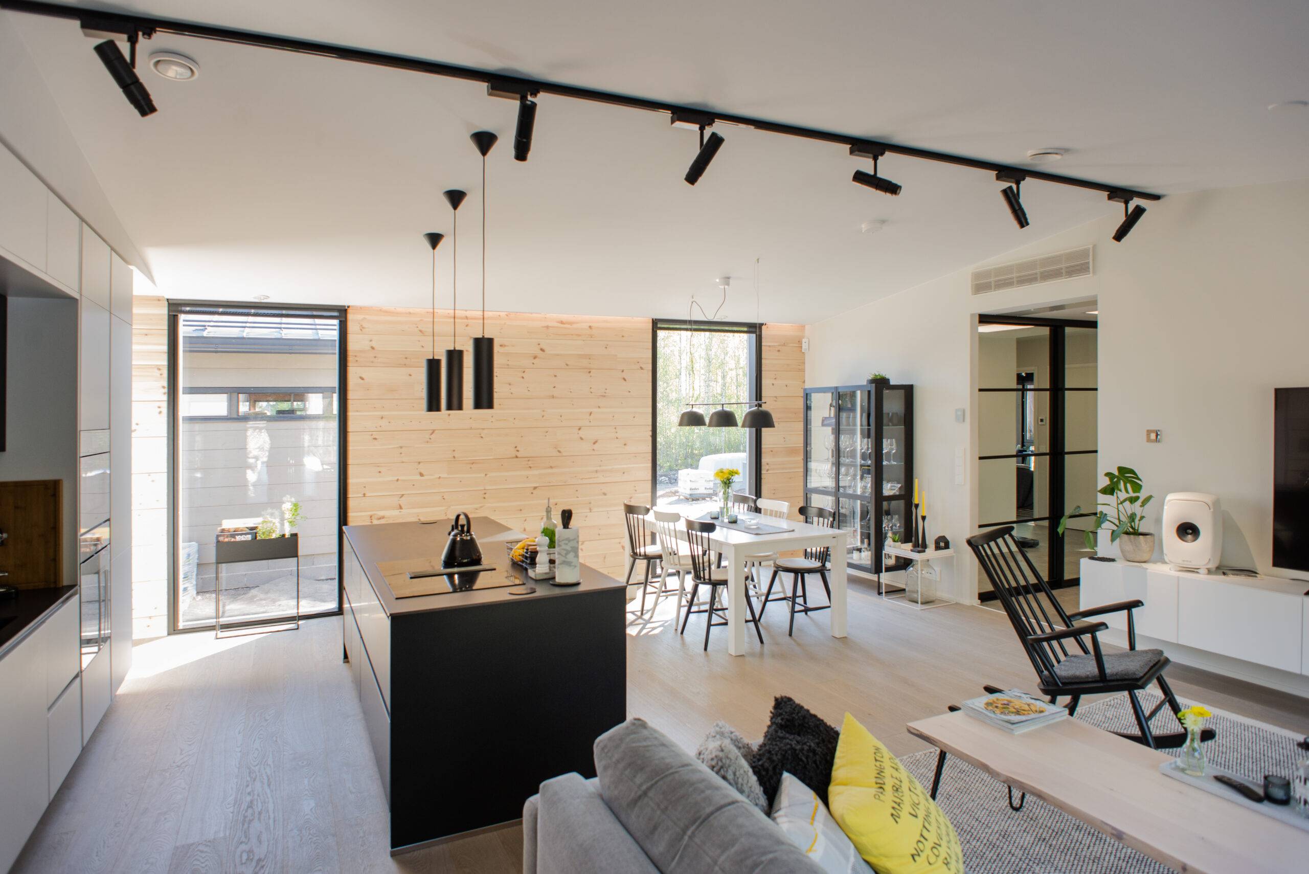 Moderna cocina y salón de planta abierta con detalles de madera y muebles negros para un aspecto elegante. La iluminación interior de alta calidad de LedStore.fi crea un gran ambiente, y los grandes ventanales dejan entrar la luz natural. 