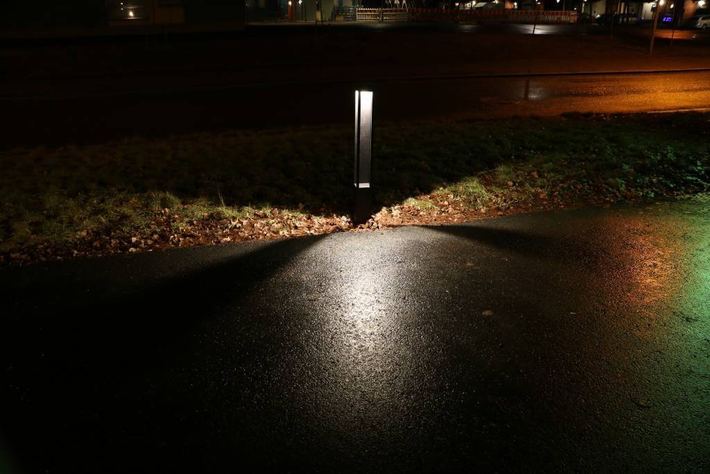 Una farola LED que ilumina una calle húmeda con patrones triangulares bordeados de hierba; un telón de fondo urbano con luces lejanas.