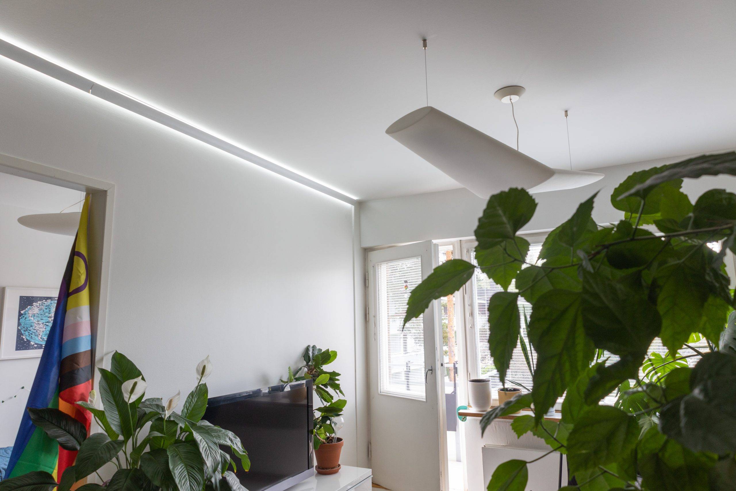 Un moderno salón de diseño minimalista cuenta con grandes plantas verdes, un televisor y una colorida bandera. En el techo hay una elegante lámpara LED junto a elegantes elementos LED, mientras que la luz natural entra a raudales por las puertas de cristal que dan al exterior.