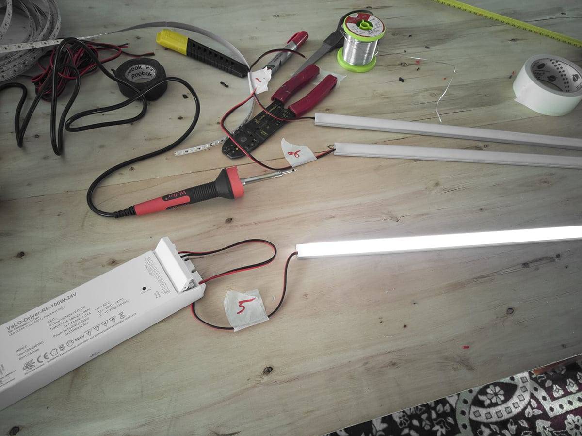 Banco de trabajo con herramientas eléctricas, tiras de LED, cables, tubos fluorescentes incandescentes; incluye alicates, multímetro, hilo de soldar y fuente de alimentación.