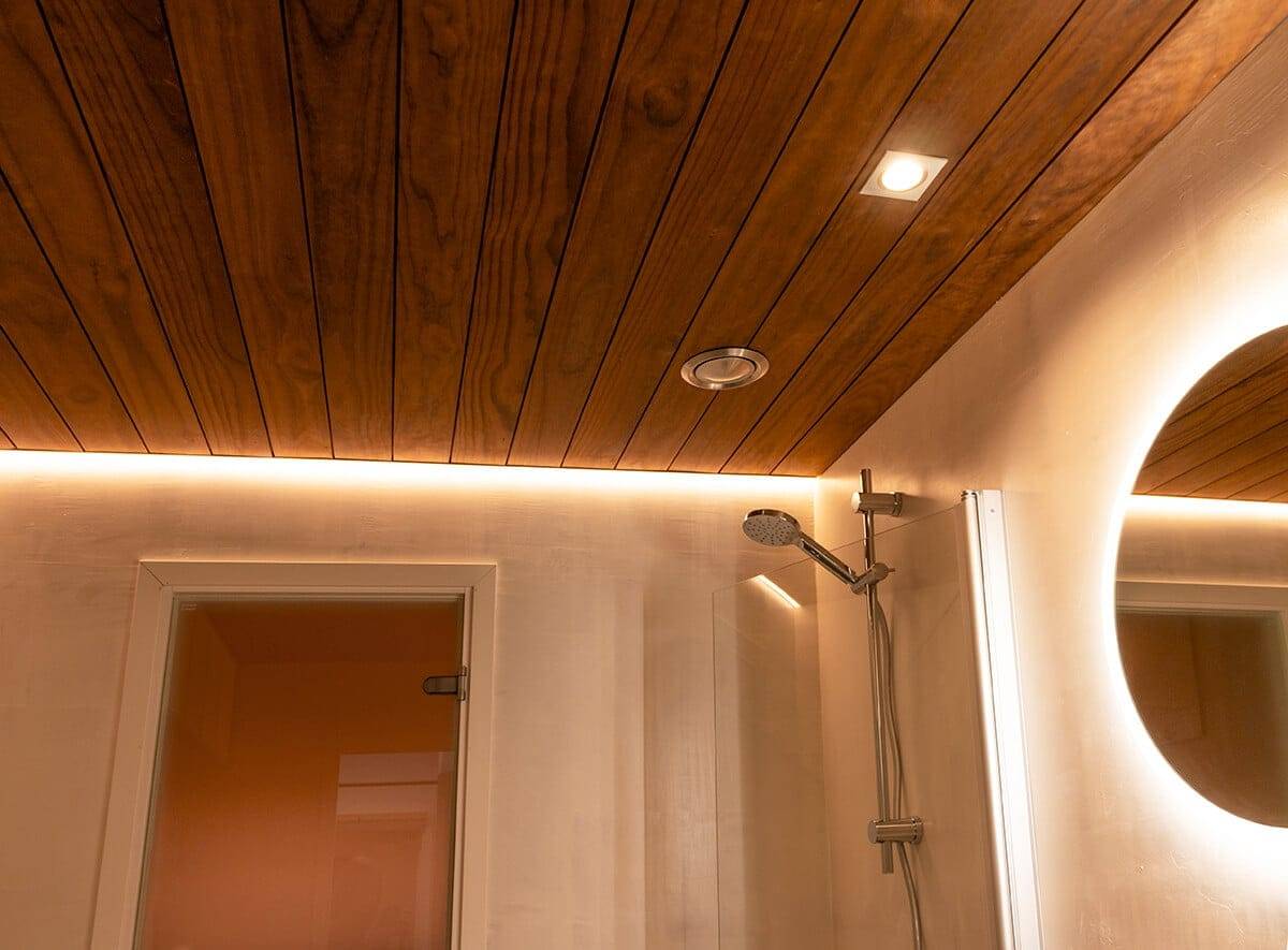 Cuarto de baño con techo de madera e iluminación LED ambiental, moderna alcachofa de ducha, luz puntual y espejo LED circular; puerta a la izquierda.