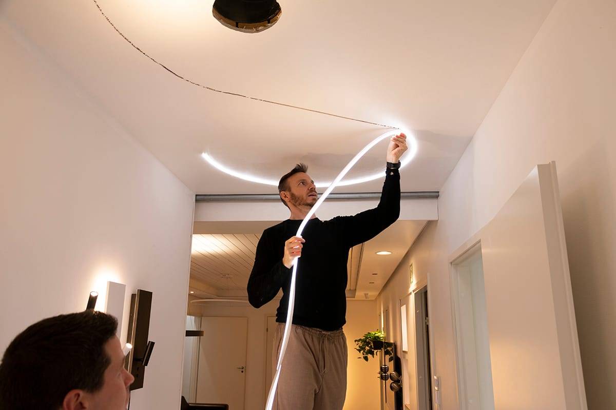 Una persona coloca una tira de luz LED flexible en un techo moderno, lo que mejora la iluminación de la habitación. Suéter negro, pantalones beige; otra persona en primer plano, borrosa.