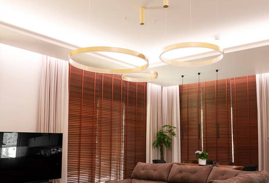 El moderno salón familiar de Kaitamäki, Espoo, cuenta con un impresionante esquema de iluminación de LedStore.fi: grandes luces anulares de color dorado crean un ambiente lujoso junto a unas persianas marrones a juego y un acogedor sofá. Rematado con un elegante televisor y una exuberante vegetación, ¡este es el tipo de iluminación que hace que tu casa sea acogedora e impresionante!
