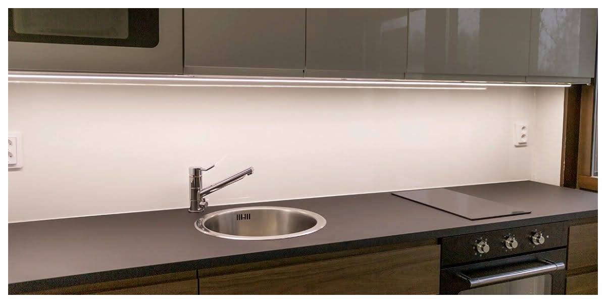 Cocina moderna con iluminación LED en los armarios, elegante encimera oscura, fregadero de acero inoxidable, grifo monomando, placa iluminada y armarios brillantes.