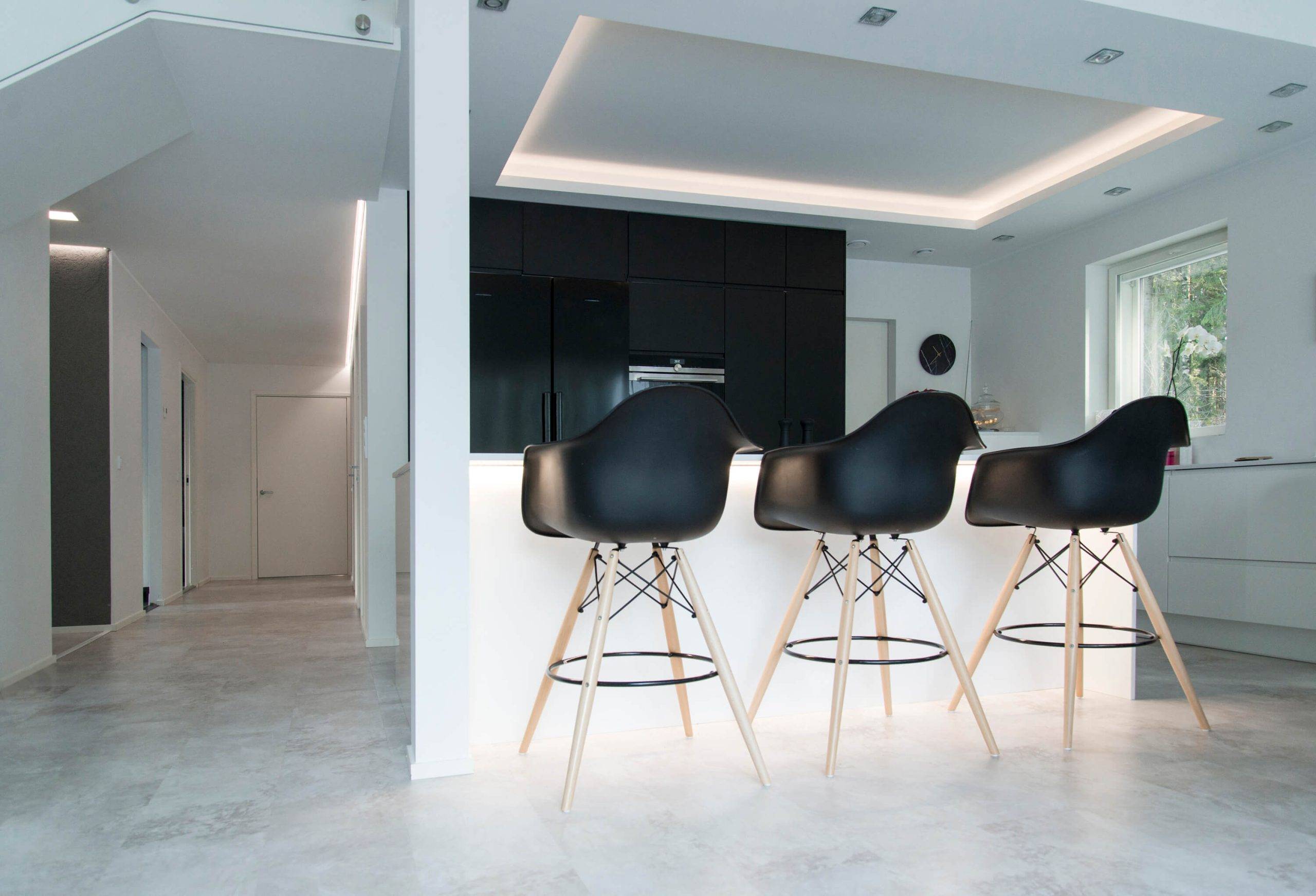 Una cocina moderna con taburetes de bar negros sobre una isla blanca, armarios oscuros, un techo empotrado con iluminación LED, una ventana y un pasillo minimalista crean un aspecto elegante.