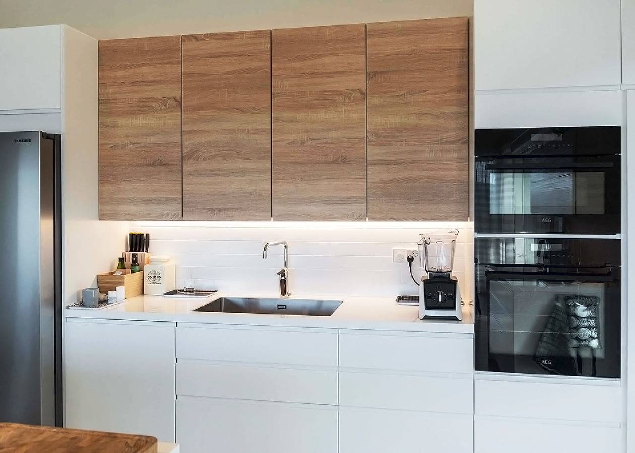 Cocina moderna con armarios superiores de madera, encimeras blancas iluminadas con LED, fregadero de acero inoxidable con grifo cromado, horno doble y electrodomésticos de encimera.