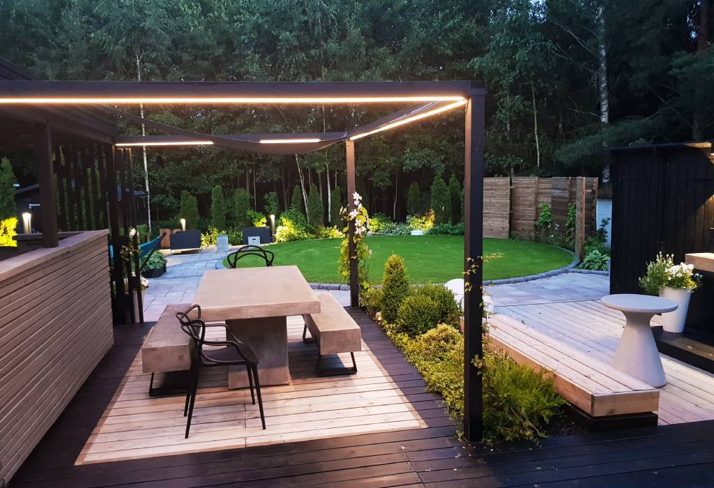 Un moderno patio trasero con una mesa de comedor de hormigón y sillas negras bajo una pérgola minimalista. La pérgola tiene iluminación LED integrada para crear una atmósfera cambiante. La zona está rodeada de vegetación verde, un único césped bien cuidado y un camino de piedra, que añaden encanto natural. Además, como pequeño detalle, hay una moderna jardinera blanca para disfrutar de la elegante terraza. Echa un vistazo a la amplia gama de iluminación LED de LedStore.fi y lleva la misma elegancia moderna a tu propio jardín.