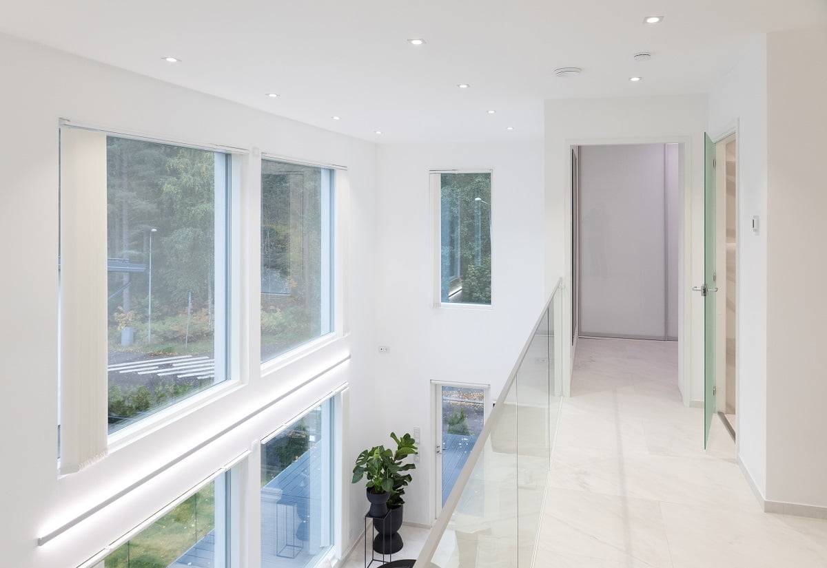 Un pasillo interior luminoso y moderno, con grandes ventanales a ambos lados, permite la entrada de luz natural. El espacio tiene paredes blancas e iluminación LED empotrada que crea un ambiente agradable para los niños. Una balaustrada de cristal añade elegancia. Una maceta verde añade un toque de color al conjunto. Bienvenido a la gama de iluminación LED de alta calidad de LedStore.fi, ¡y crea el espacio luminoso de tus sueños!