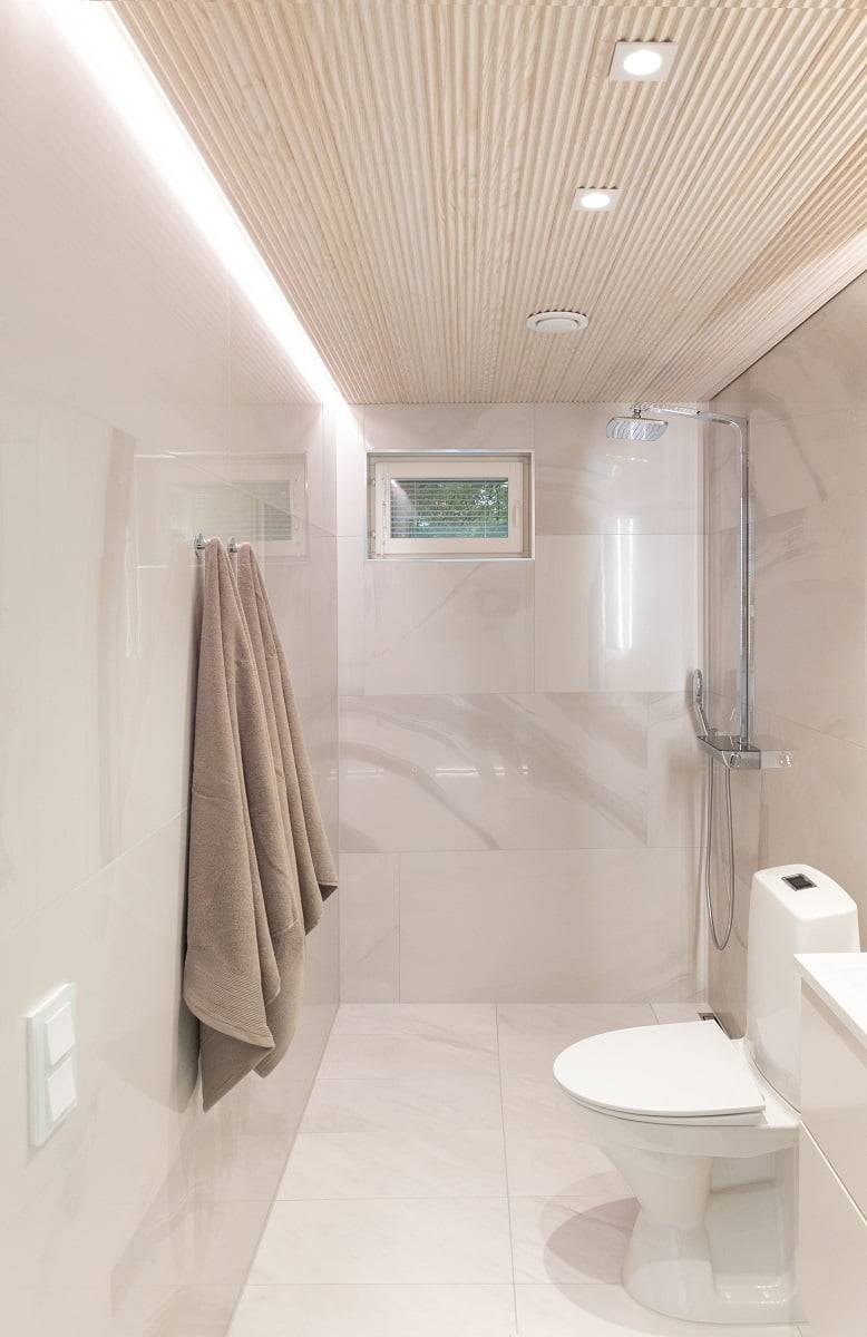 Un cuarto de baño moderno y minimalista, con paredes de color beige claro y techo de baldosas de madera. Un toallero de pared con dos toallas, una pequeña ventana y una zona de ducha separada por una mampara de cristal añaden elegancia al espacio. Las luces LED integradas en el techo proporcionan una iluminación eficaz. LedStore.fi: Descubre soluciones de iluminación LED perfectas para interiores elegantes y atemporales.