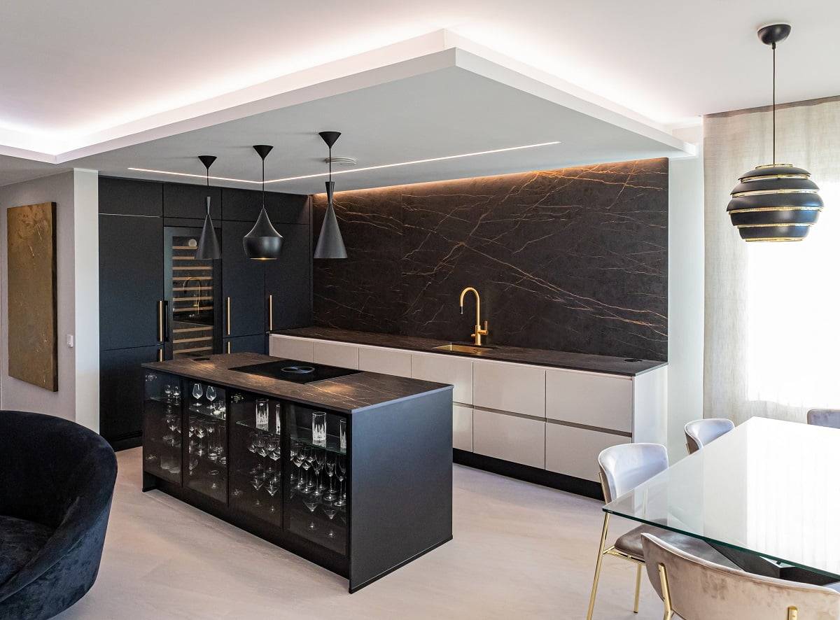 Una cocina moderna con azulejos blancos y negros, baldosas blancas, una encimera de mármol negro y un grifo dorado. Unas lámparas colgantes iluminan una isla negra con cristalería con fugas. Una mesa de comedor adyacente de cristal tiene sillas tapizadas y sobre ella brilla una llamativa lámpara colgante de LED iluminada en negro y dorado. Echa un vistazo a la amplia gama de iluminación LED de LedStore.fi para crear un aspecto impresionante similar en tu propia casa.