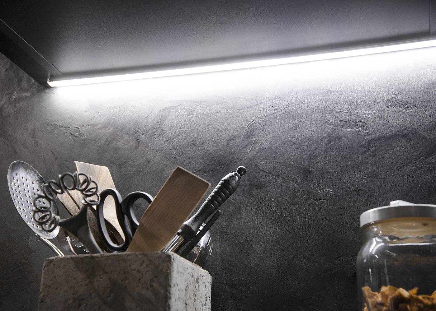 Foto de cocina con vajilla en un estante, una tira de luz LED brillante resalta la pared oscura texturizada; a la derecha se ve un tarro de cristal. Ideal para iluminación LED moderna.