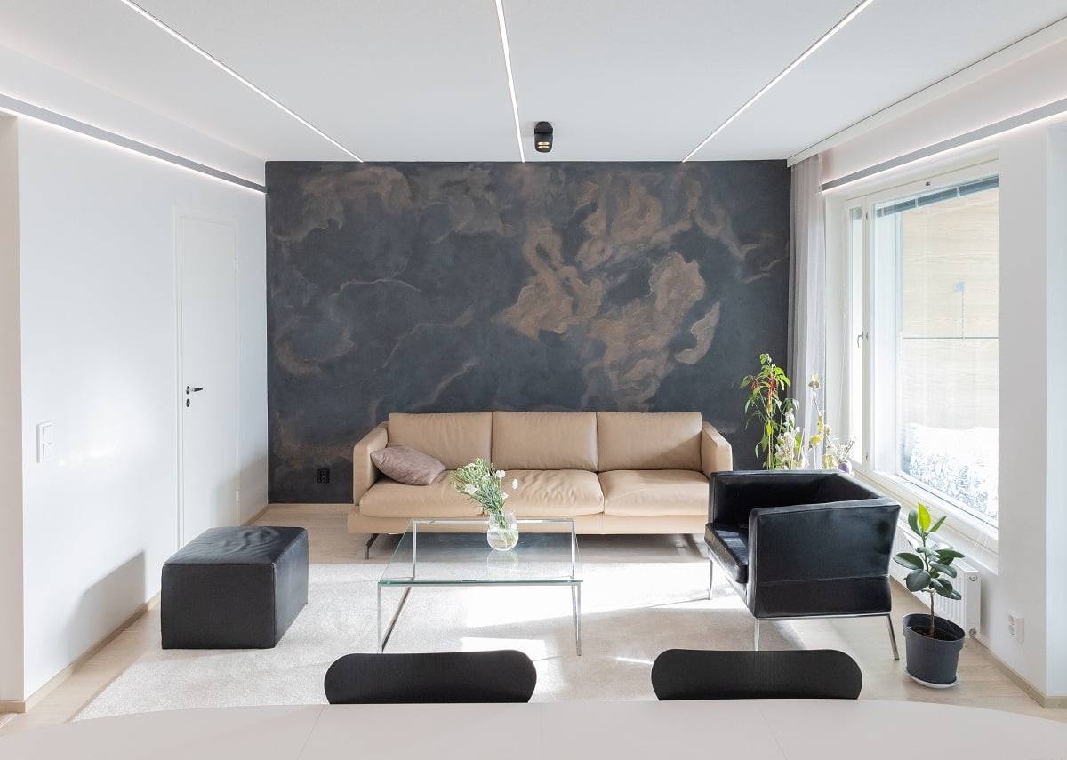 El moderno salón tiene un sofá beige, un sillón negro y una mesa de centro de cristal, que hacen juego con la pared de efecto oscuro. Junto a la ventana hay una planta verde cerca de una pequeña paja negra. Encarga un foco, complementado con luces LED salvajes en LedStore.fi.