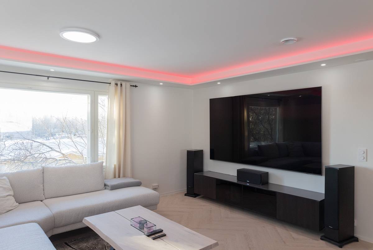 Un salón moderno lleno de ambiente elegante. Un gran sofá te invita a relajarte, mientras que una mesa de centro de madera clara añade calidez al espacio. Un televisor de pantalla plana montado en la pared con retroiluminación LED añade un toque moderno a la habitación. Amplifica tu experiencia con altavoces de suelo y disfruta de la iluminación de acento LED roja del techo. Una gran ventana deja entrar la luz natural, haciendo que el espacio parezca amplio y luminoso. Todos estos elementos se combinan para crear el ambiente perfecto con las versátiles soluciones de iluminación LED de LedStore.fi.