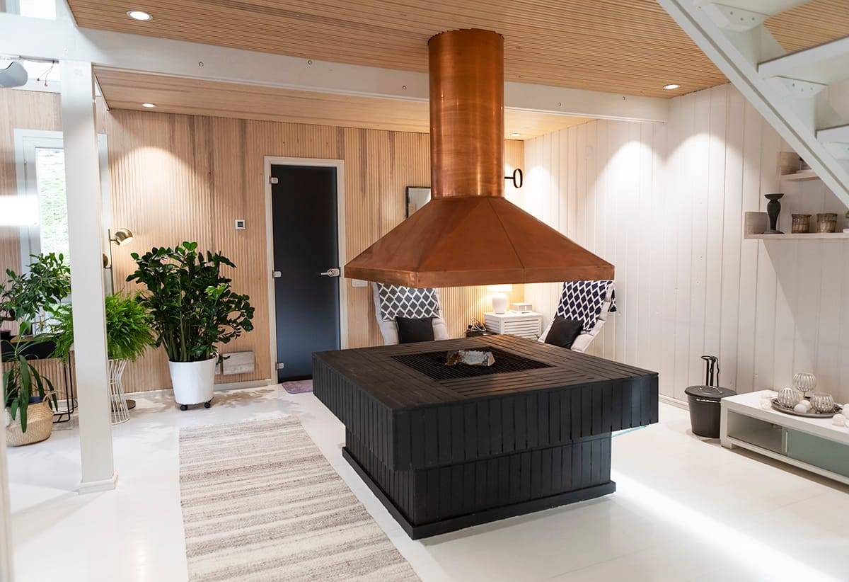 Salón moderno con chimenea negra y de madera en el centro, tubo de cobre, iluminación LED, sillas tapizadas, vegetación y decoración elegante.