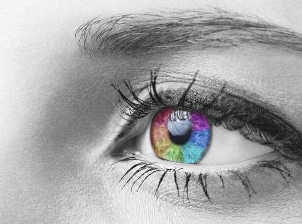 Primer plano de un ojo en tonos grises con iris iridiscente en colores brillantes: rojo, azul, verde, morado; fuerte contraste con la piel y las pestañas monocromas.