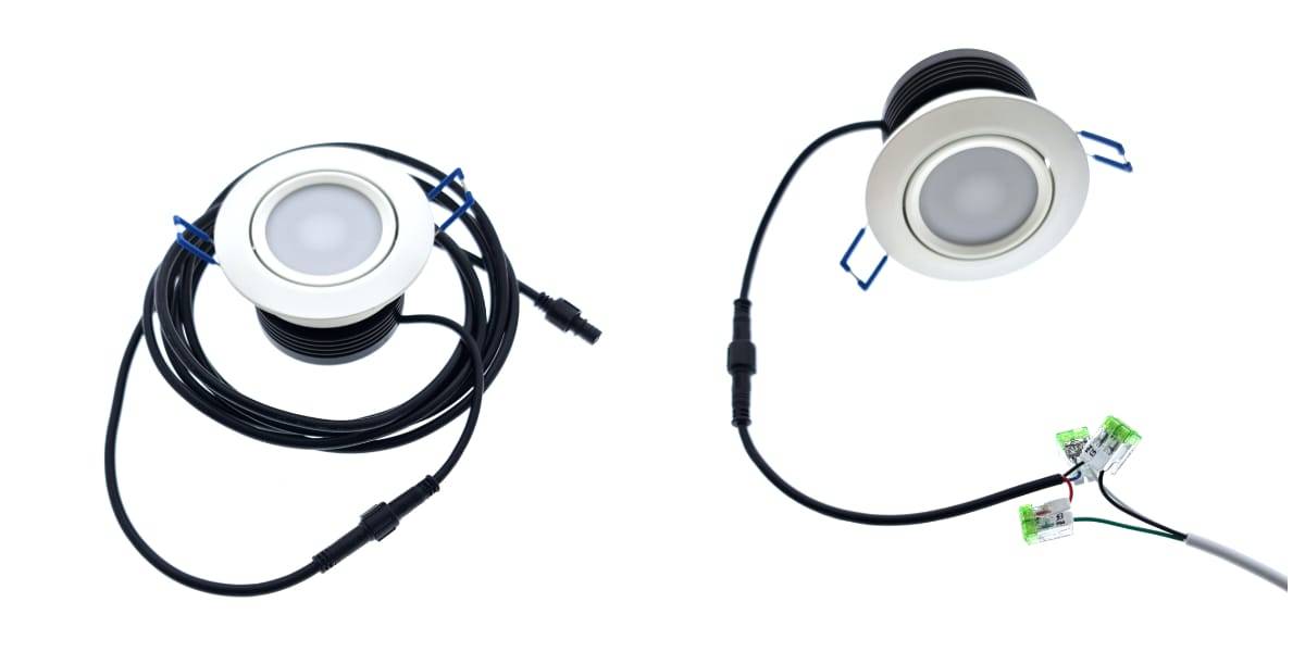 Dos downlights LED redondos con marco blanco están unidos a largos cables negros. Las luces tienen conectores en los extremos de los cables. Un cable está trenzado, mientras que el otro se extiende y conecta a los conectores verdes en línea, destacando su propia y elegante integración LED.