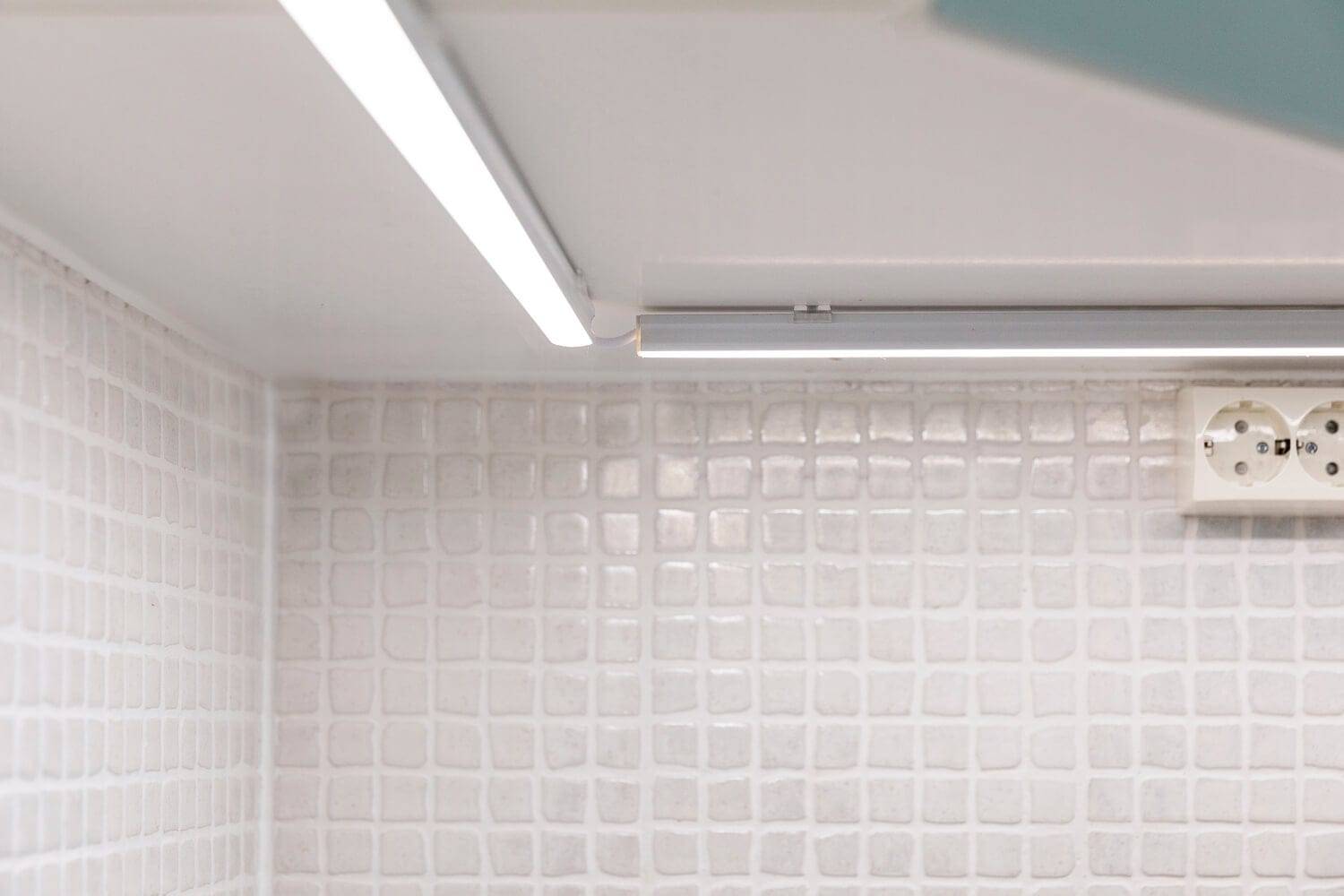 Un moderno salpicadero de cocina con azulejos blancos texturizados, iluminado por una luz LED lineal situada debajo del armario; un suave resplandor resalta la toma de corriente de la pared.